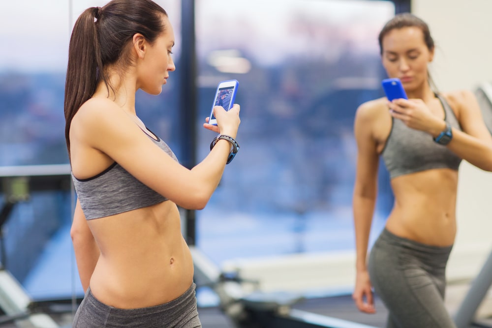 sport, fitness, lifestyle, technology and people concept - young woman with smartphone taking mirror selfie in gym